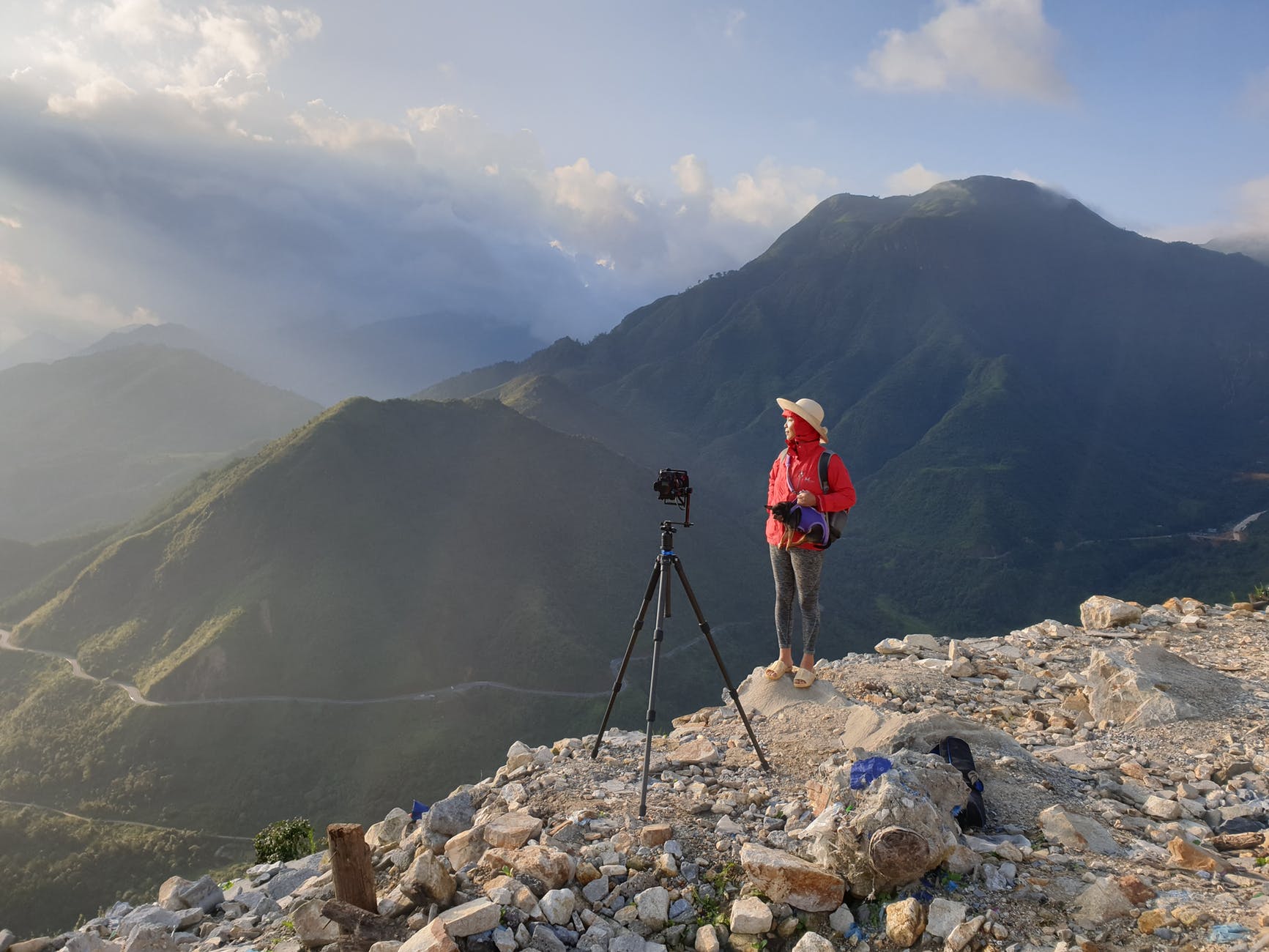 person wearing red jacket standing on cliff beside black camera tripod